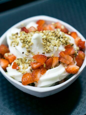 A white ceramic bowl on a dark table. The bowl contains yogurt, sliced strawberries, and chopped nuts. Photo by Fábio Alves on Unsplash