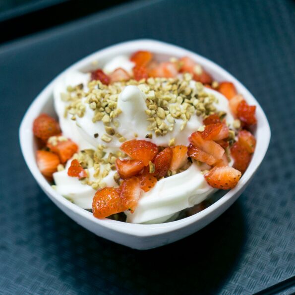 A white ceramic bowl on a dark table. The bowl contains yogurt, sliced strawberries, and chopped nuts. Photo by Fábio Alves on Unsplash