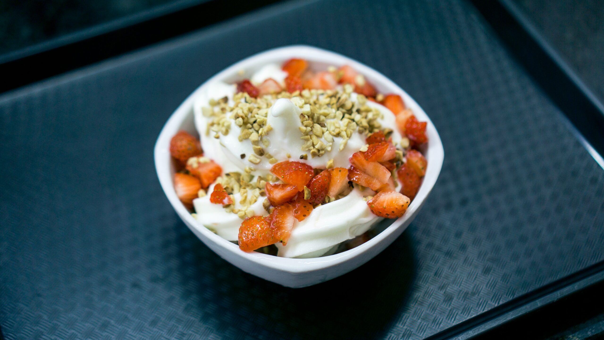 A white ceramic bowl on a dark table. The bowl contains yogurt, sliced strawberries, and chopped nuts. Photo by Fábio Alves on Unsplash