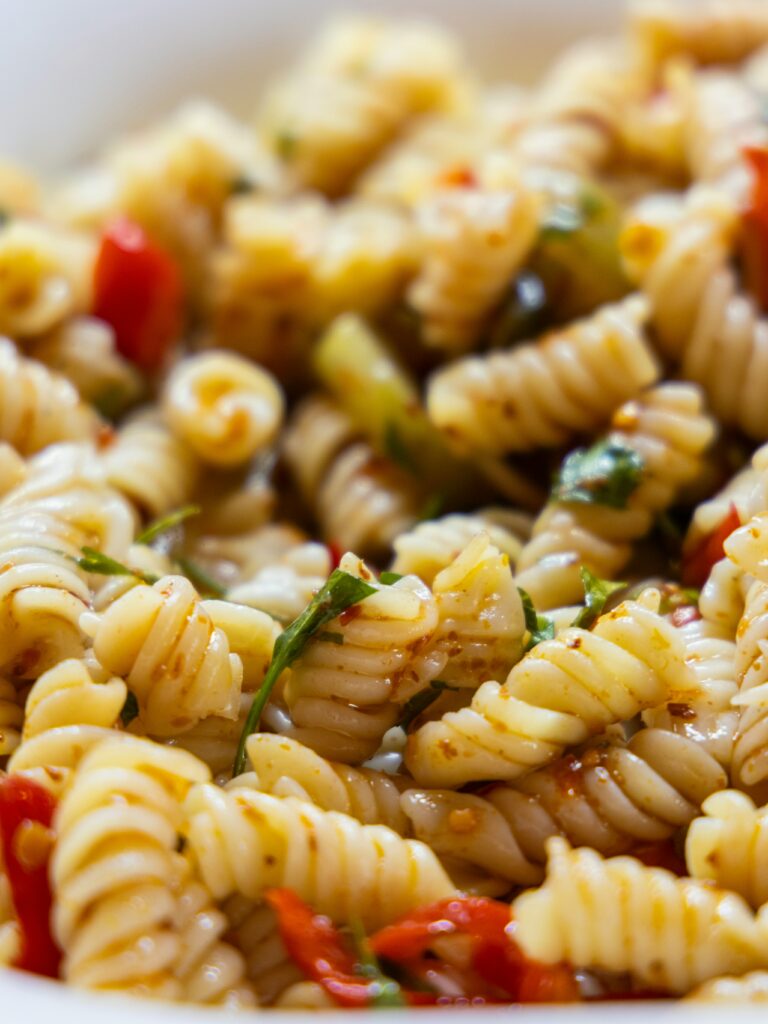 A white bowl contains spiral pasta, diced vegetables, and a vinaigrette dressing. Photo by engin akyurt on Unsplash.
