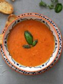 A white bowl on a dark surface. The bowl has a patterned rim and contains tomato soup garnished with a sprig of basil. The table contains crumbs and toasted bread. Photo by Julia Kicova on Unsplash
