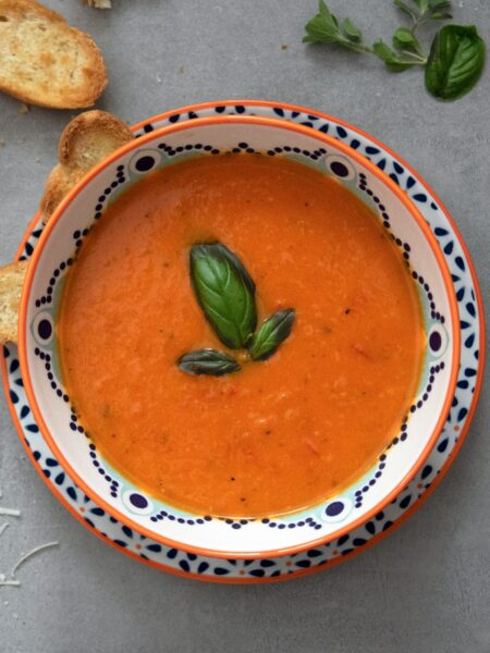 A white bowl on a dark surface. The bowl has a patterned rim and contains tomato soup garnished with a sprig of basil. The table contains crumbs and toasted bread. Photo by Julia Kicova on Unsplash