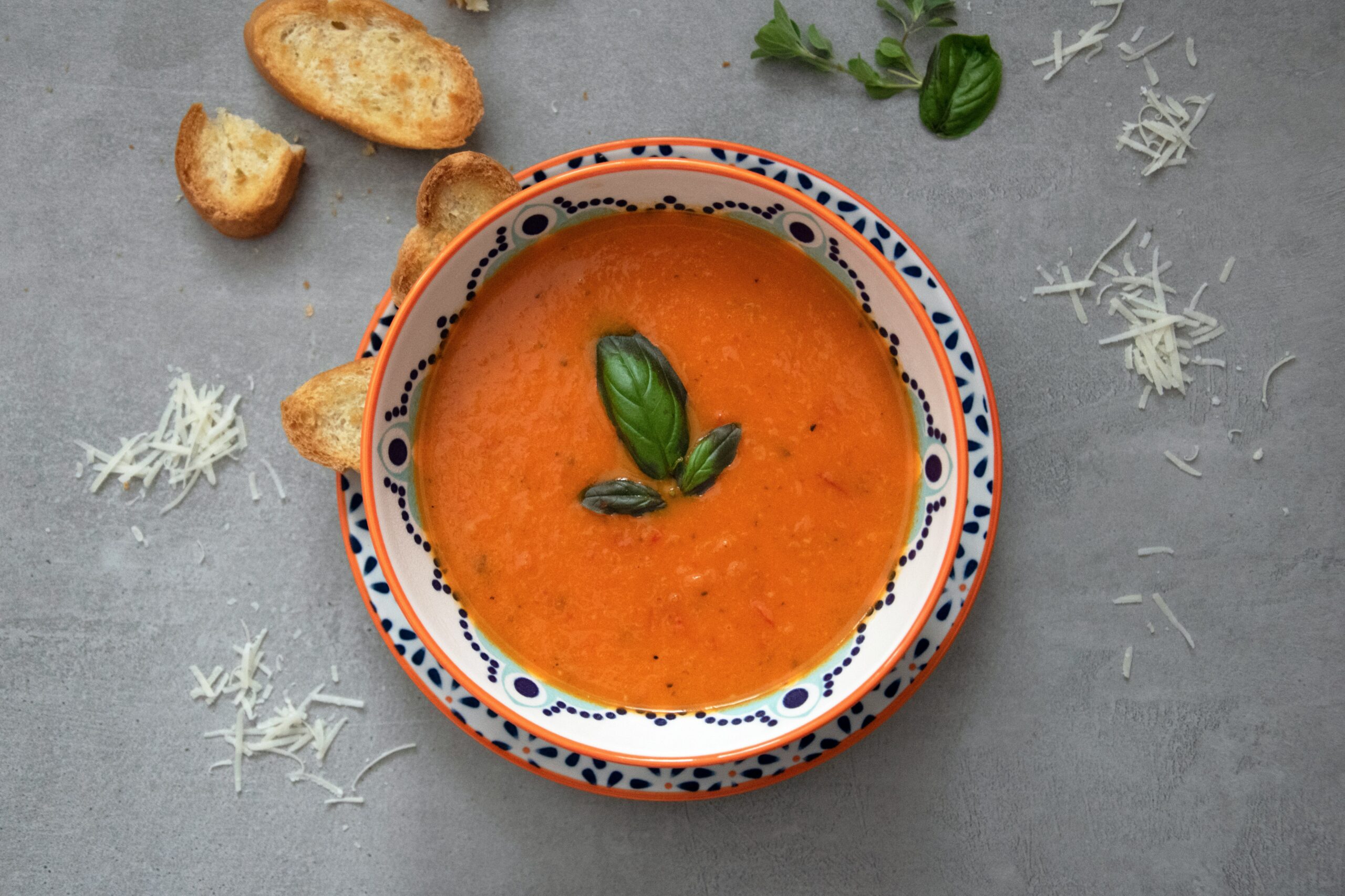 A white bowl on a dark surface. The bowl has a patterned rim and contains tomato soup garnished with a sprig of basil. The table contains crumbs and toasted bread. Photo by Julia Kicova on Unsplash