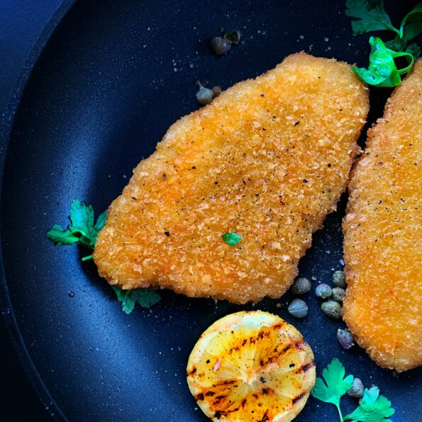 A dark blue bowl contains a piece of baked or fried chicken, a grilled lemon slice, some capers, and some sprigs of cilantro. Photo by LikeMeat on Unsplash