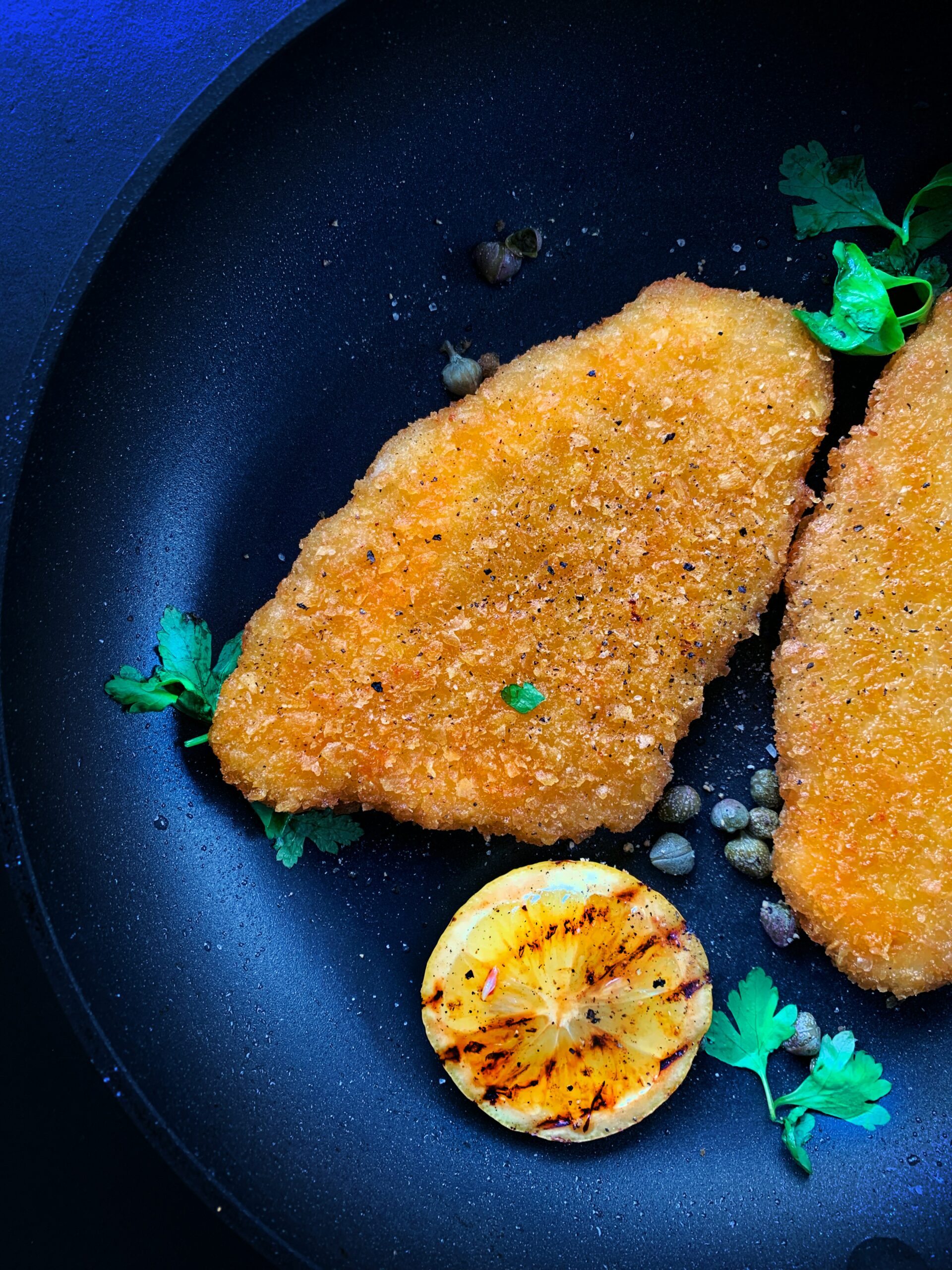 A dark blue bowl contains a piece of baked or fried chicken, a grilled lemon slice, some capers, and some sprigs of cilantro. Photo by LikeMeat on Unsplash