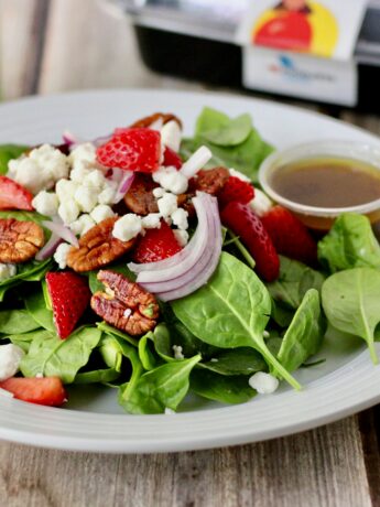 A white ceramic plate rests on a wooden table along with a napkin, knife, fork, to-go container, and a green can of La Croix sparkling water. On the plate is a salad composed of spinach leaves, pecan pieces, red onion slices, strawberry pieces, goat cheese, and a small plastic container of vinaigrette dressing. Photo by logan jeffrey on Unsplash.