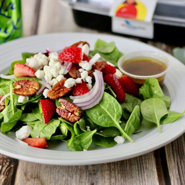 A white ceramic plate rests on a wooden table along with a napkin, knife, fork, to-go container, and a green can of La Croix sparkling water. On the plate is a salad composed of spinach leaves, pecan pieces, red onion slices, strawberry pieces, goat cheese, and a small plastic container of vinaigrette dressing. Photo by logan jeffrey on Unsplash.