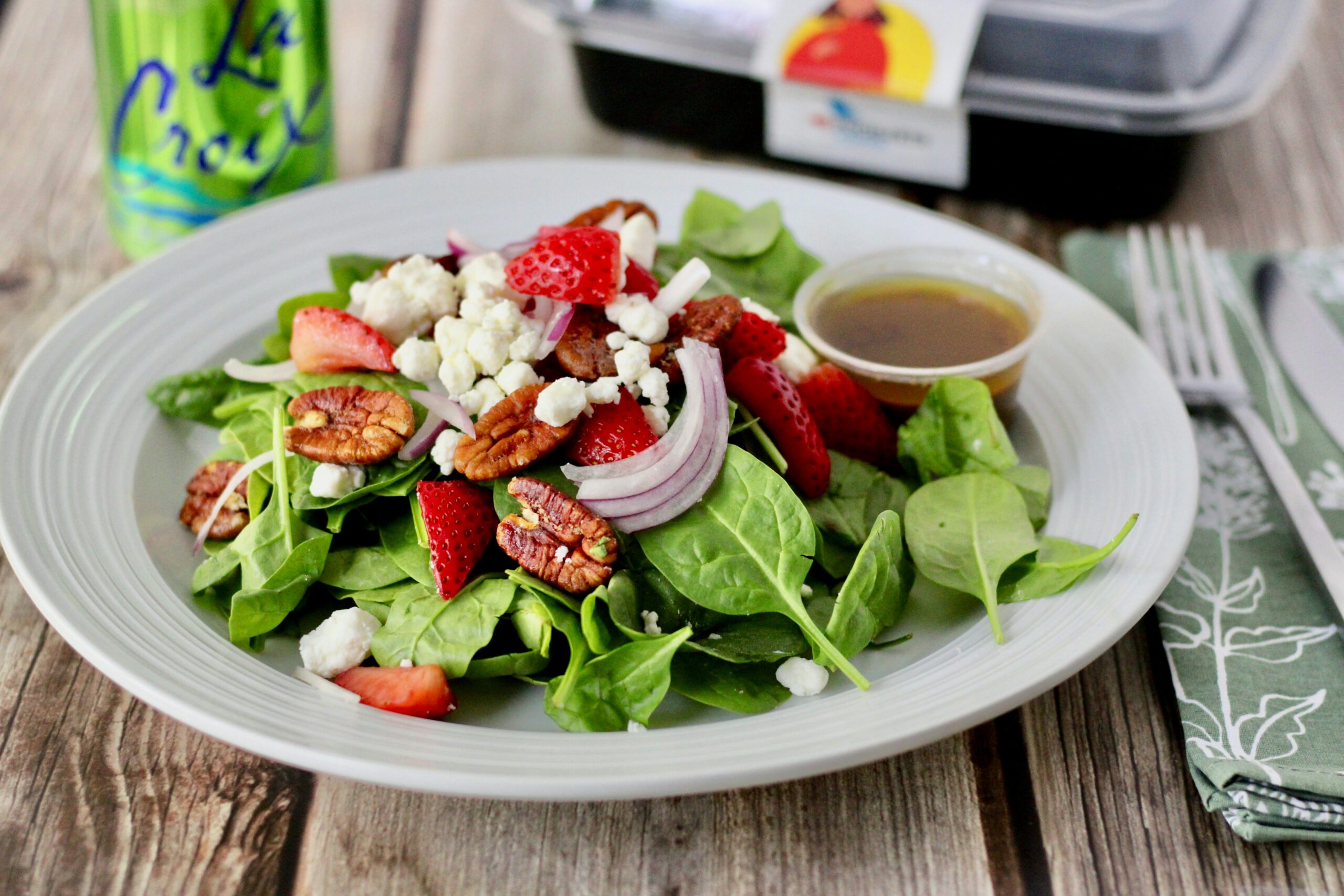 A white ceramic plate rests on a wooden table along with a napkin, knife, fork, to-go container, and a green can of La Croix sparkling water. On the plate is a salad composed of spinach leaves, pecan pieces, red onion slices, strawberry pieces, goat cheese, and a small plastic container of vinaigrette dressing. Photo by logan jeffrey on Unsplash.