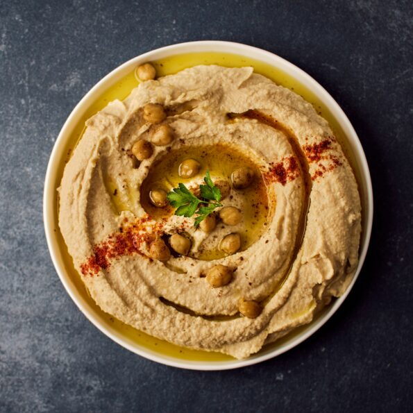 White bowl on a dark table. Inside the bowl is hummus garnished with chickpeas and herbs. Photo by Ludovic Avice on Unsplash