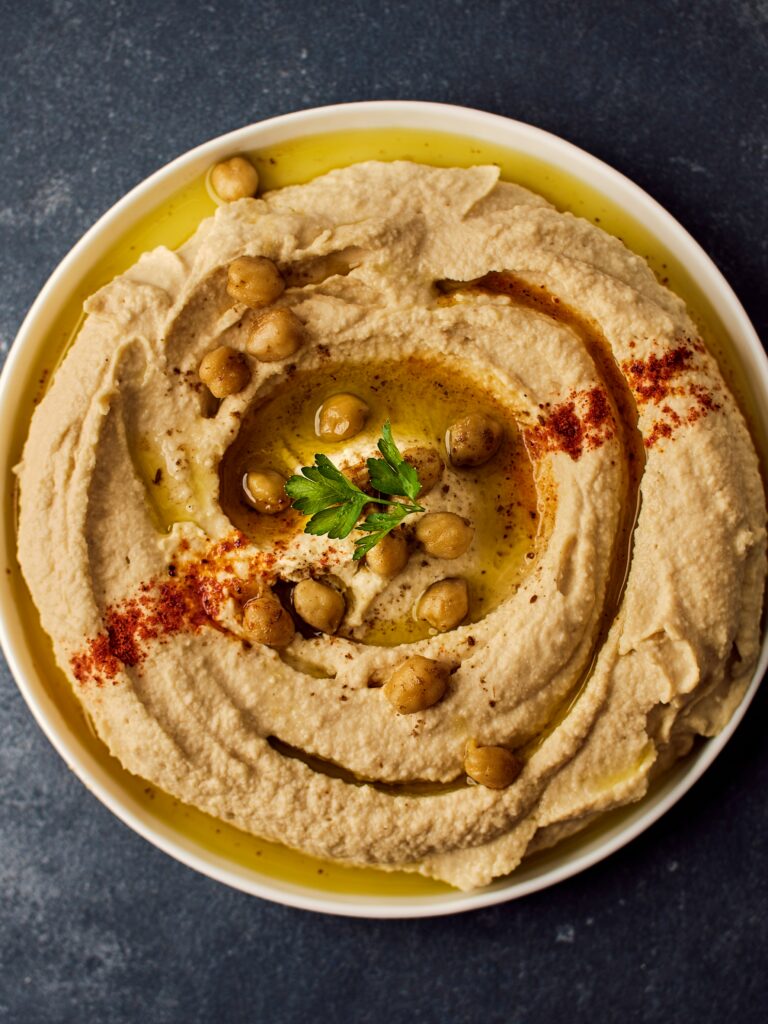 White bowl on a dark table. Inside the bowl is hummus garnished with chickpeas and herbs. Photo by Ludovic Avice on Unsplash