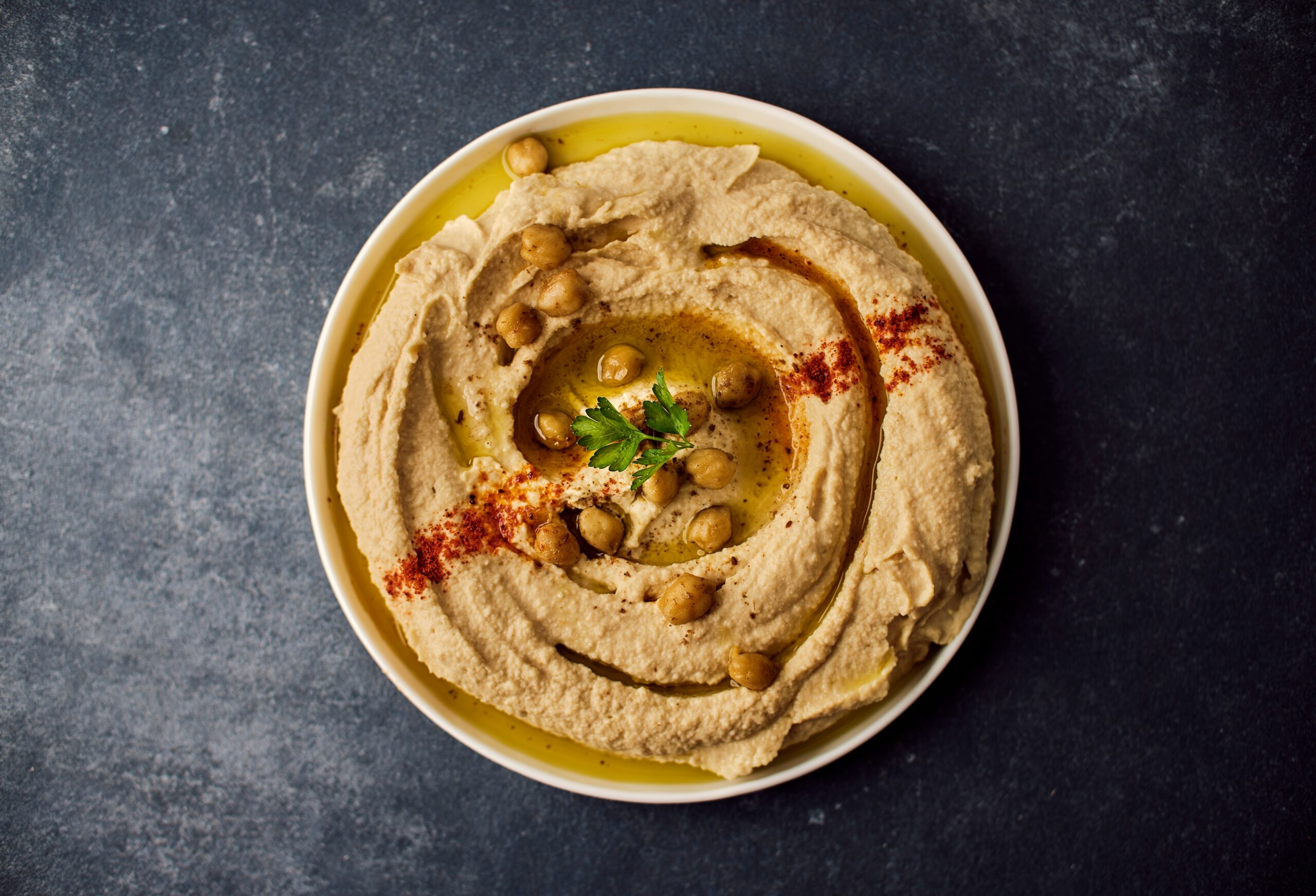 White bowl on a dark table. Inside the bowl is hummus garnished with chickpeas and herbs. Photo by Ludovic Avice on Unsplash