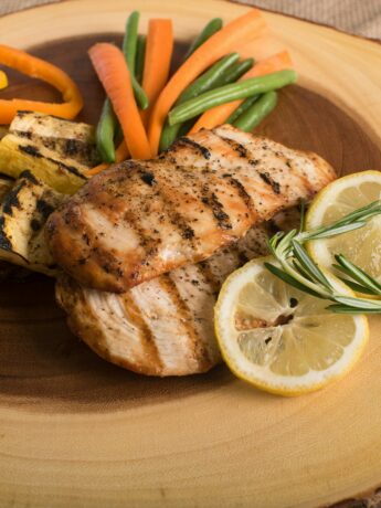 A sliced piece of log (wood) serves as a plate. On the plate are grilled squash, bell pepper slides, carrots, green beans, grilled chicken breasts, and two slices of lemon with a sprig of rosemary. Photo by Mark DeYoung on Unsplash