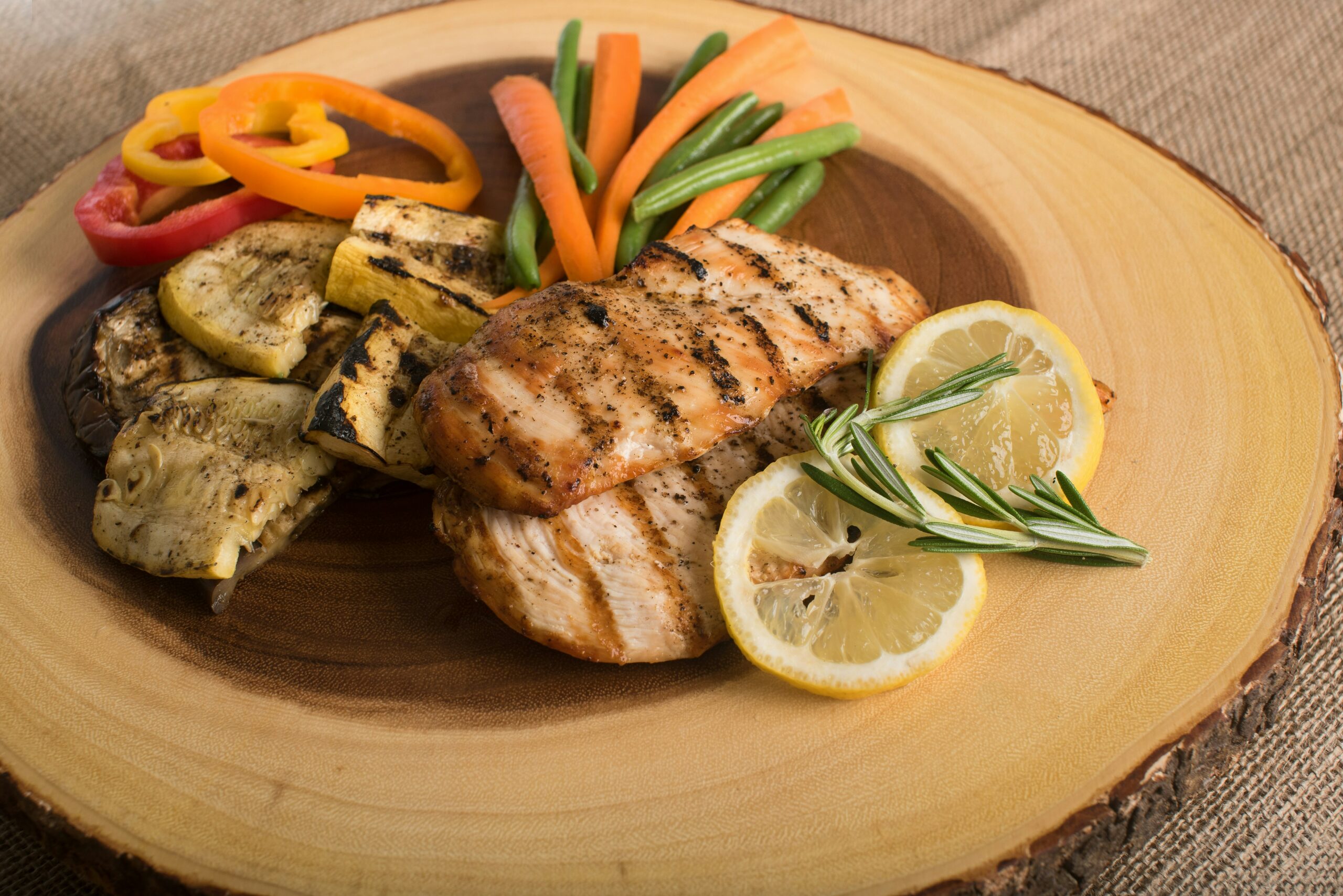A sliced piece of log (wood) serves as a plate. On the plate are grilled squash, bell pepper slides, carrots, green beans, grilled chicken breasts, and two slices of lemon with a sprig of rosemary. Photo by Mark DeYoung on Unsplash