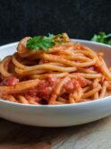 White ceramic bowl of pasta with red sauce, garnished with basil. ON the table are some tomatoes and herbs. Photo by Pratik Gupta on Unsplash