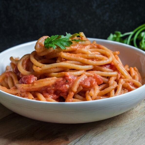 White ceramic bowl of pasta with red sauce, garnished with basil. ON the table are some tomatoes and herbs. Photo by Pratik Gupta on Unsplash
