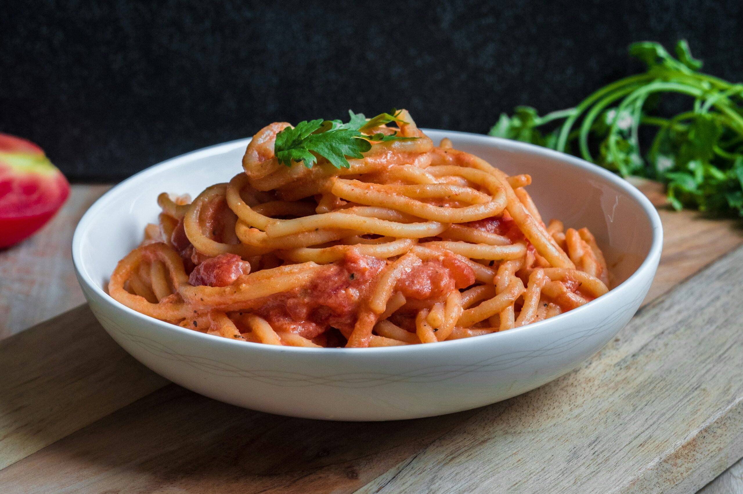 White ceramic bowl of pasta with red sauce, garnished with basil. ON the table are some tomatoes and herbs. Photo by Pratik Gupta on Unsplash