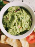 White ceramic bowl n a wooden surface. The bowl contains bright green guacamole. Also on the table are tomatoes, limes, cilantro. Photo by Tessa Rampersad on Unsplash