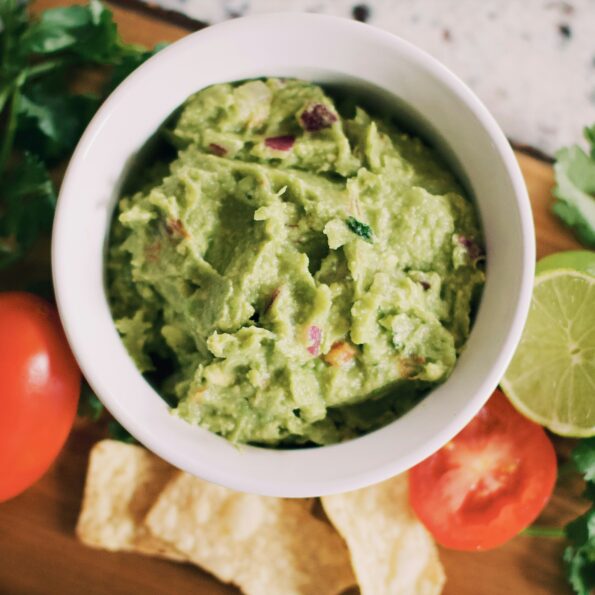 White ceramic bowl n a wooden surface. The bowl contains bright green guacamole. Also on the table are tomatoes, limes, cilantro. Photo by Tessa Rampersad on Unsplash