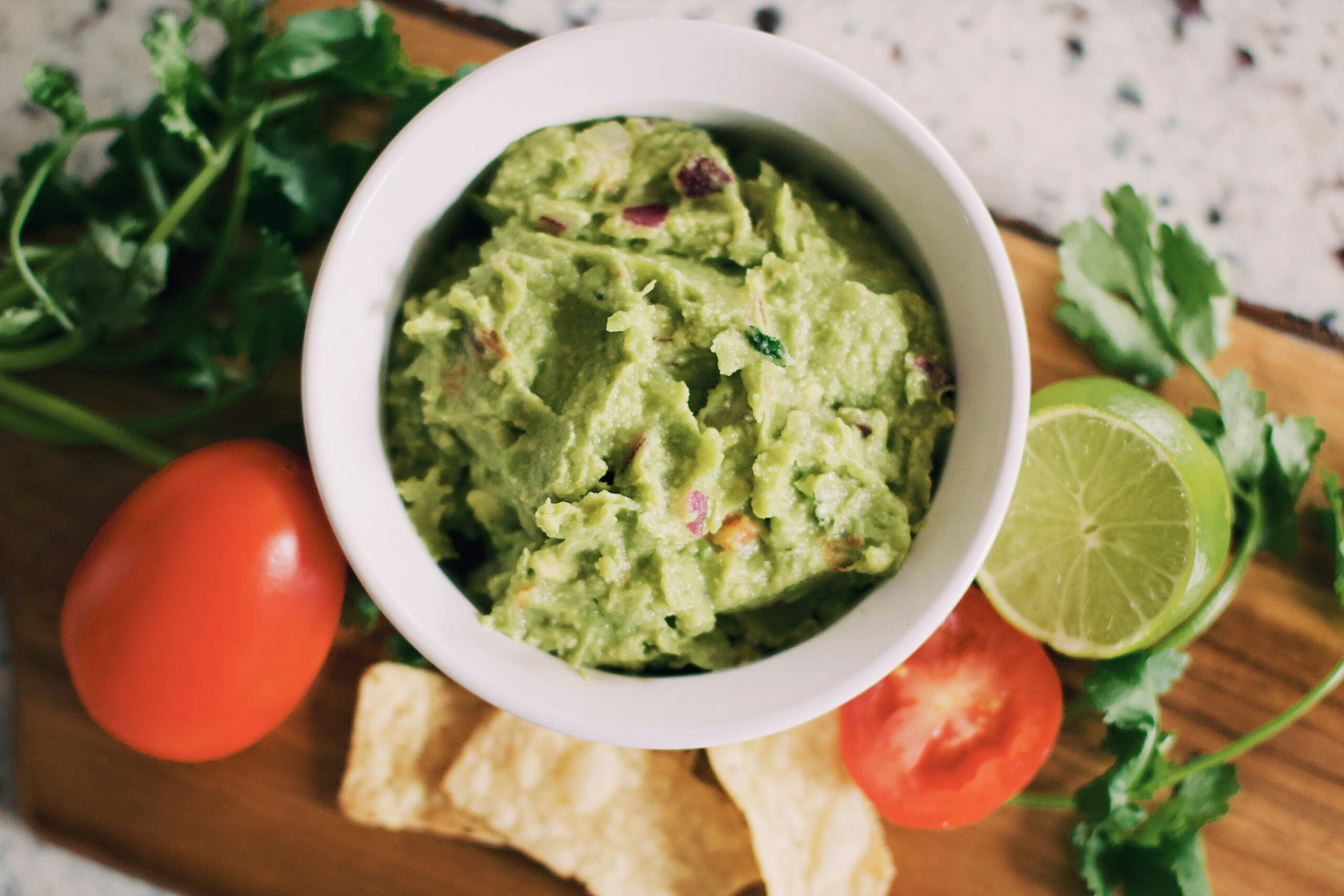 White ceramic bowl n a wooden surface. The bowl contains bright green guacamole. Also on the table are tomatoes, limes, cilantro. Photo by Tessa Rampersad on Unsplash